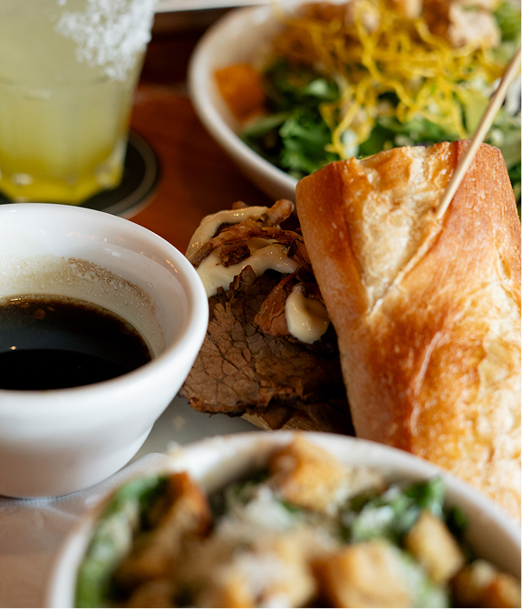 Close-up of a beef dip sandwich with au jus, salad, and a cocktail at Warehouse.