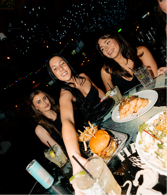 Group of friends enjoying burgers, share plates, and cocktails at a Warehouse table.