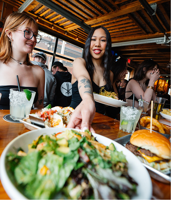 Friends sharing burgers, salads, tacos, and drinks at a Warehouse table.