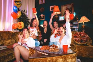 Group of friends cheering with drinks and eating wings on a retro couch during a lively night at Warehouse Ontario.