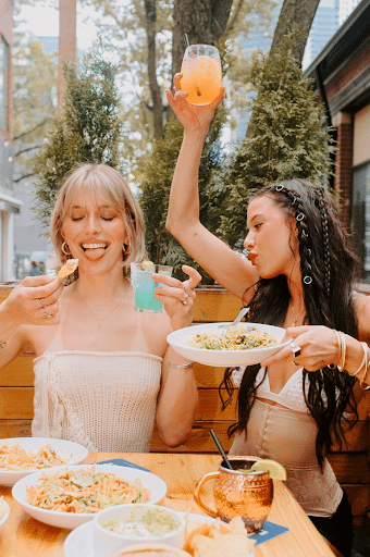 Two women toasting with vibrant cocktails and enjoying salads on Warehouse's outdoor patio in Ontario.