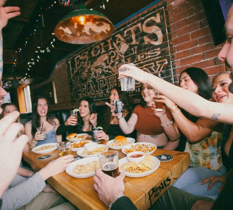 Friends raising drinks around a table with burgers, fries, mac & cheese, and nachos under warm lighting at Warehouse Whistler.