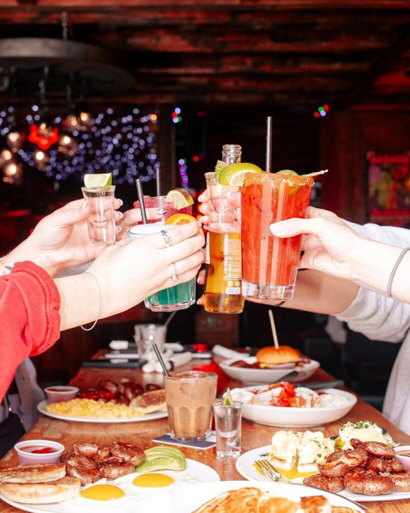 Friends clinking cocktails, beer, and shots over a brunch table at Warehouse with bright natural light.