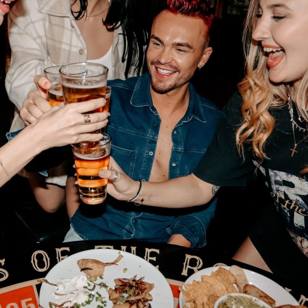 Group of friends at Warehouse Ontario toast with pints of beer, laughing around a shared meal table.