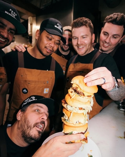 Kitchen crew celebrating over a towering stack of juicy burgers at Warehouse, laughing and admiring their creation.