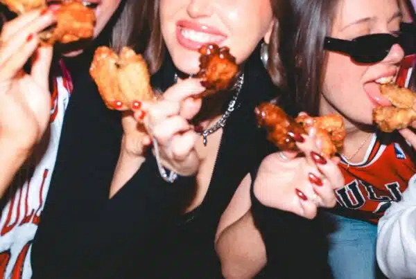 Three friends smiling and holding plates of saucy BBQ chicken wings inside a lively Warehouse bar in Toronto.