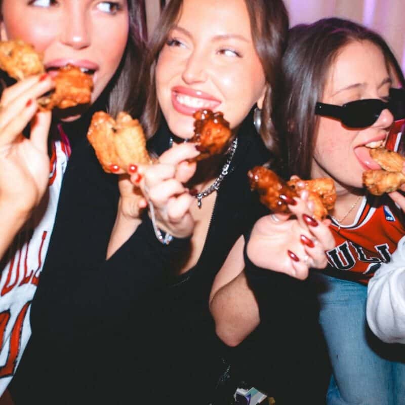 Three friends smiling and holding plates of saucy BBQ chicken wings inside a lively Warehouse bar in Toronto.