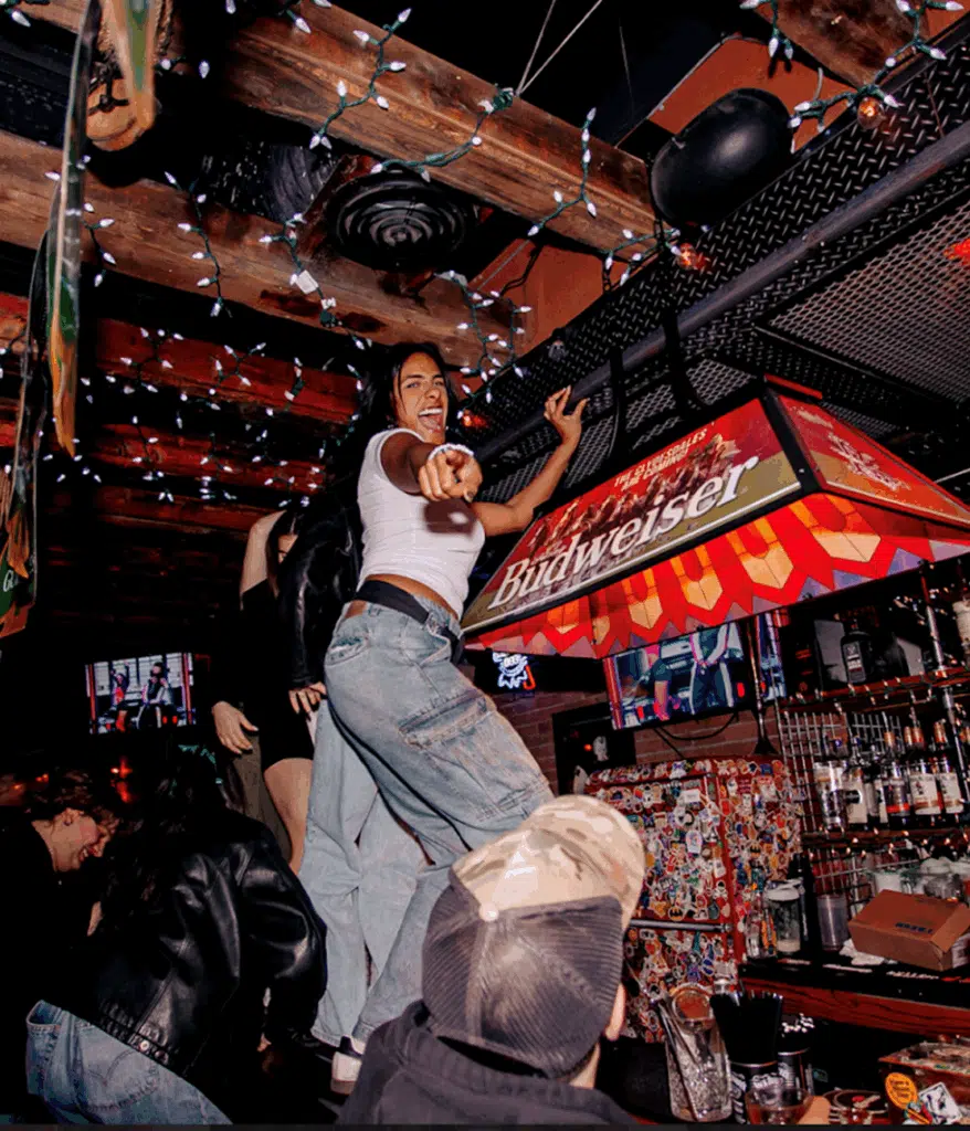 Woman dancing on the bar under string lights at Warehouse, pointing playfully as the crowd cheers her on