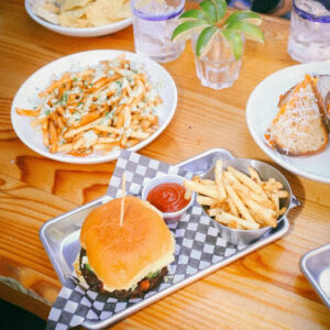 Stacked burger, golden fries, loaded cheesy fries, and grilled cheese served on wood table at El Furniture Warehouse Vancouver.