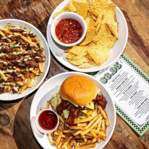 Overhead shot of bacon cheeseburger with fries, loaded fries with jalapeño sauce, and chips with salsa beside the $6.95 Happy Hour Menu.