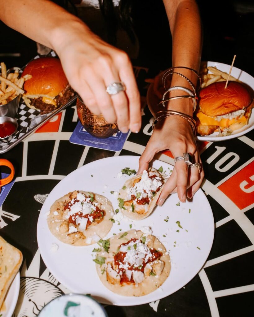 Hand garnishing tacos beside burgers and colorful cocktails on a table at El Furniture Warehouse Vancouver.