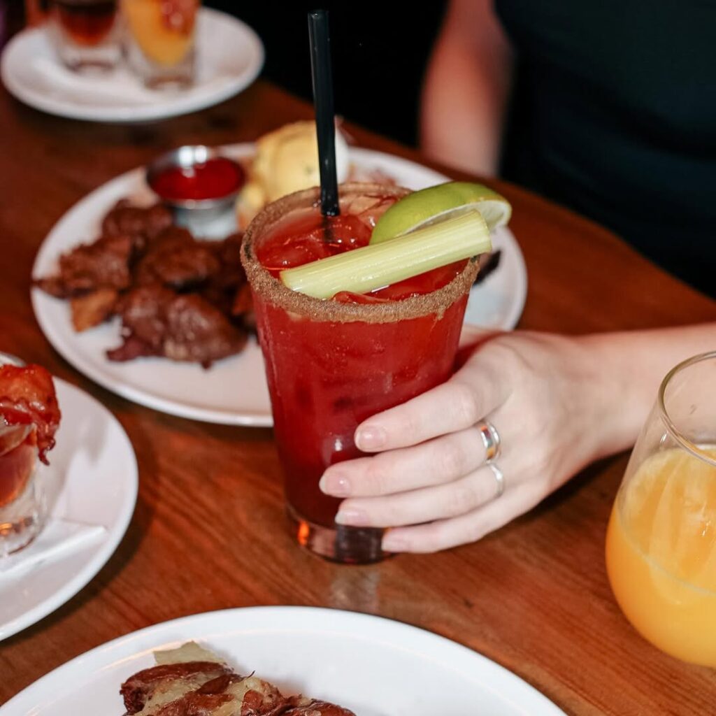 Close-up of a Caesar cocktail garnished with celery and lime, surrounded by happy hour food on a wooden table.
