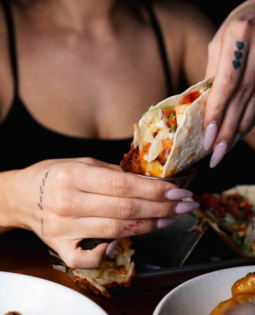 Close-up of a woman dipping a crispy fish taco filled with slaw, tomato, and cheese into creamy sauce.