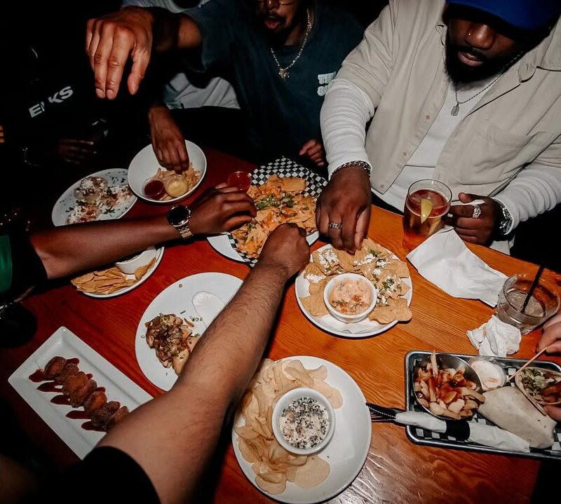 Group of friends sharing nachos, tacos, chips, and dips at a lively table during Warehouse weekend brunch.