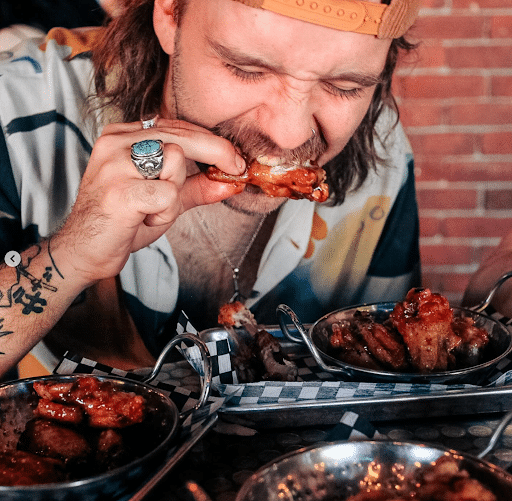 Man in a backward cap digs into saucy chicken wings at Warehouse, surrounded by metal trays of affordable food.