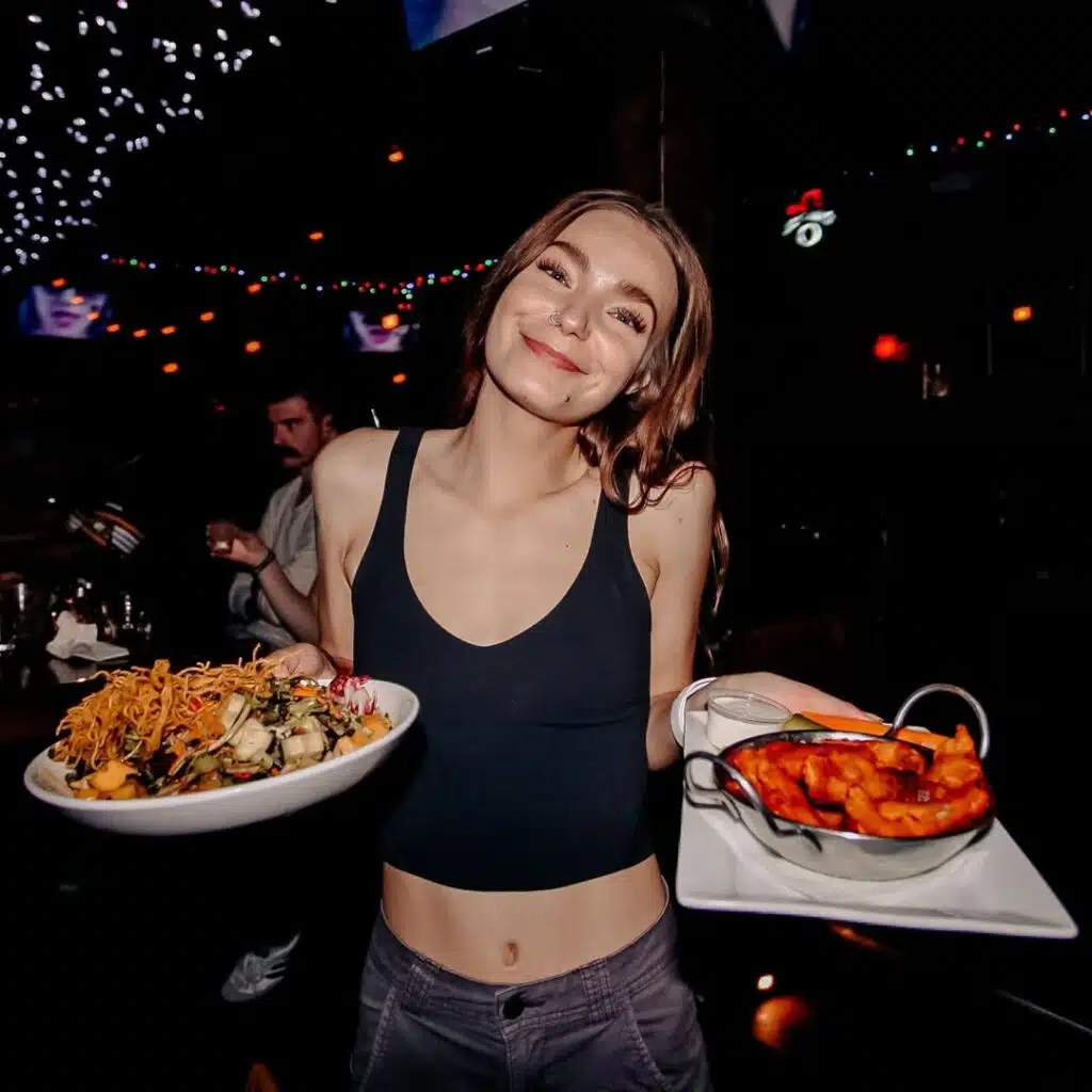 Smiling Warehouse server holding an Asian Mango Salad and a basket of crispy wings, lit by colourful string lights.