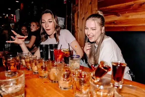 Two women laughing at a lively bar table filled with shot glasses and cocktails during Warehouse happy hour.