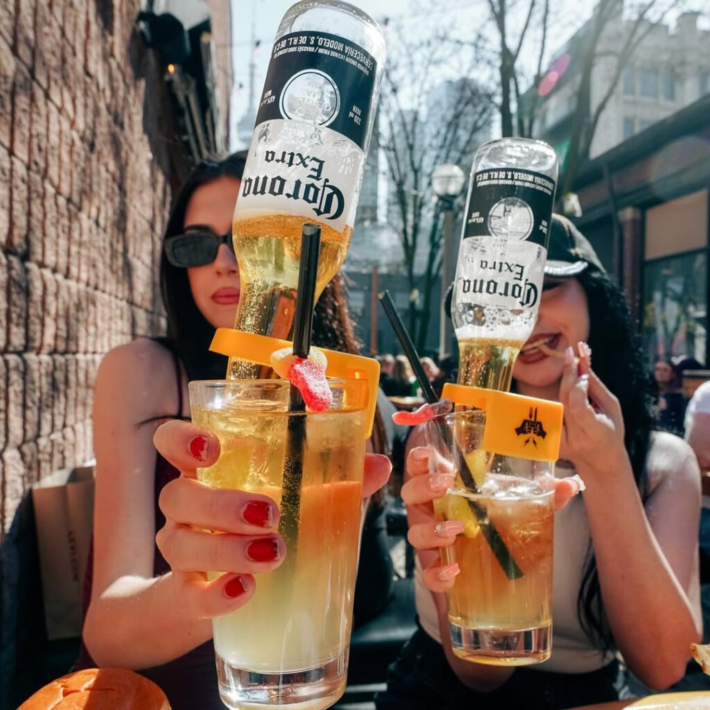 Two friends toasting with Key-Rona cocktails, featuring Corona bottles inverted into fruity patio drinks with candy garnish.