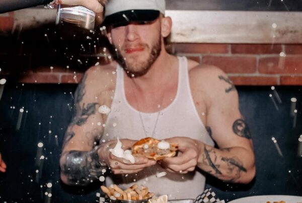 Man in a white tank top eating at a table with beer sprayed mid-air, surrounded by fries, nachos, and shots at Warehouse.