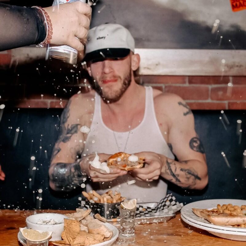 Man in a white tank top eating at a table with beer sprayed mid-air, surrounded by fries, nachos, and shots at Warehouse.
