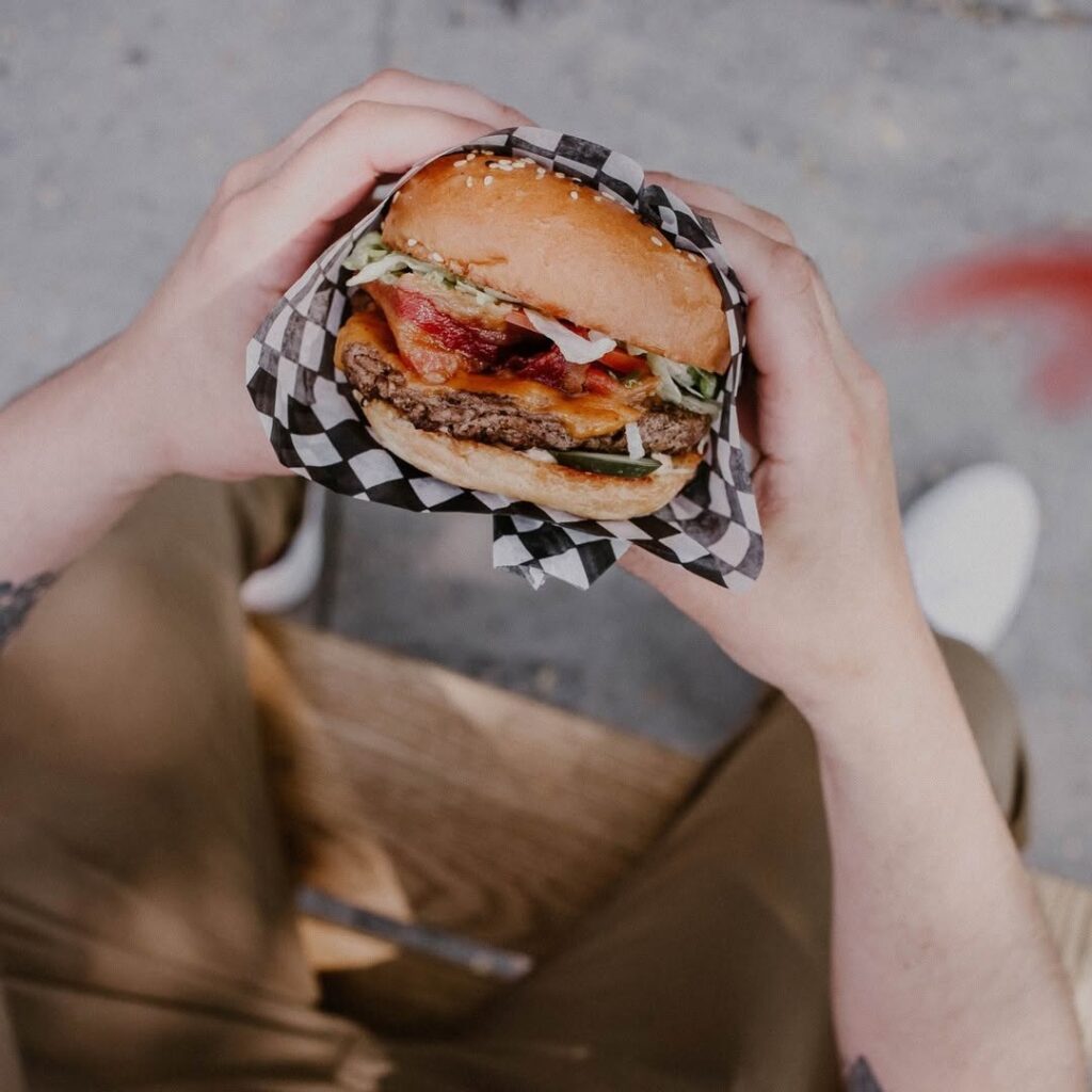 Close-up of a person holding a stacked bacon cheeseburger wrapped in checkered paper at Warehouse Ottawa.