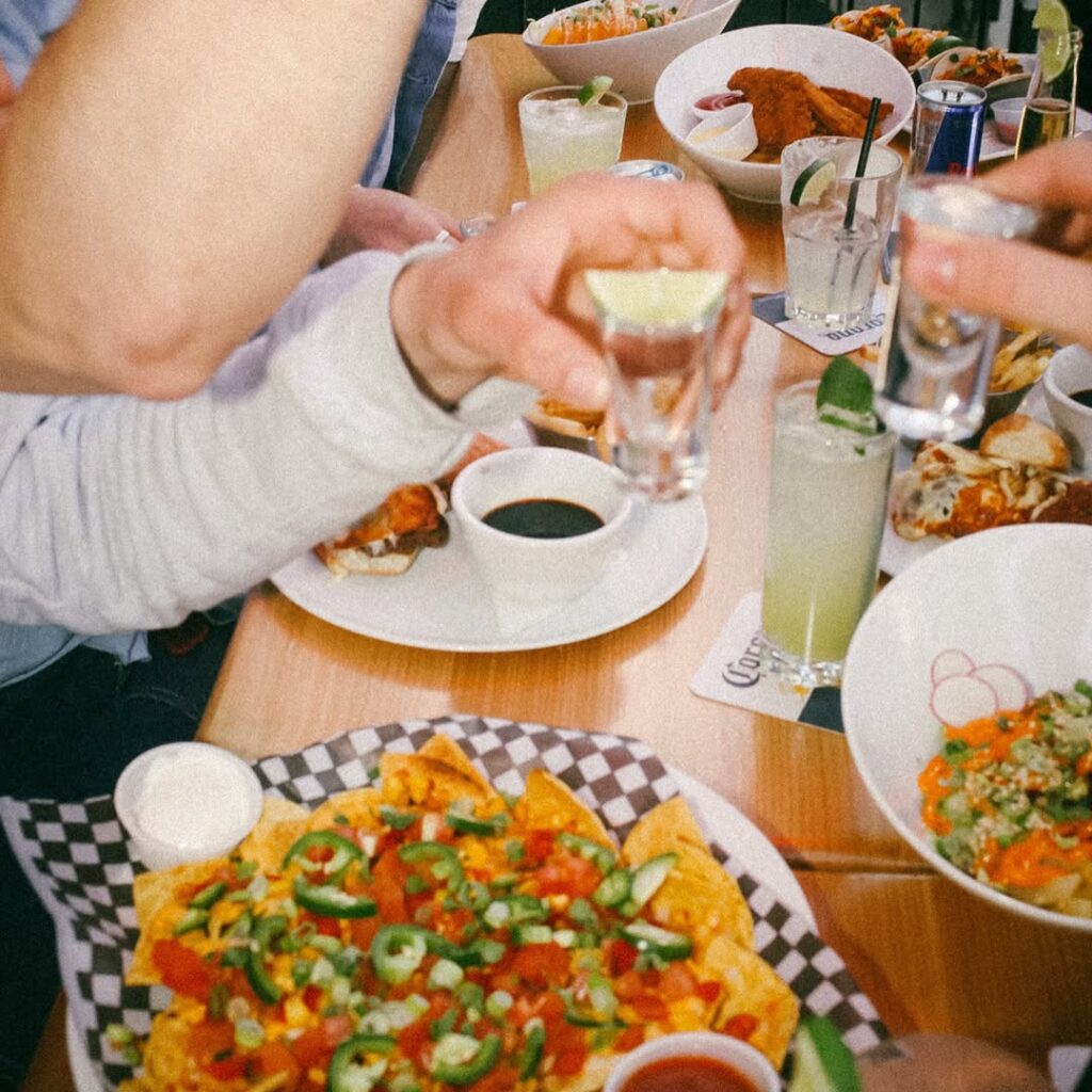 Group toasting with tequila shots over a table filled with nachos, tacos, and cocktails at Warehouse.