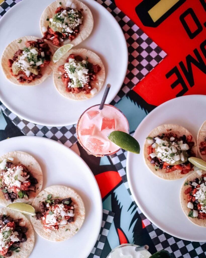 Overhead shot of assorted street tacos with lime wedges and a pink cocktail on a bold patterned table at Warehouse.