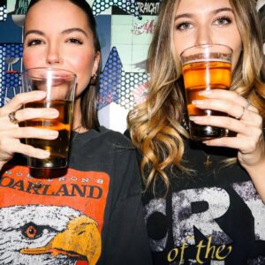 Two women in graphic tees holding up pint glasses of beer with a comic-style mural in the background at Warehouse.