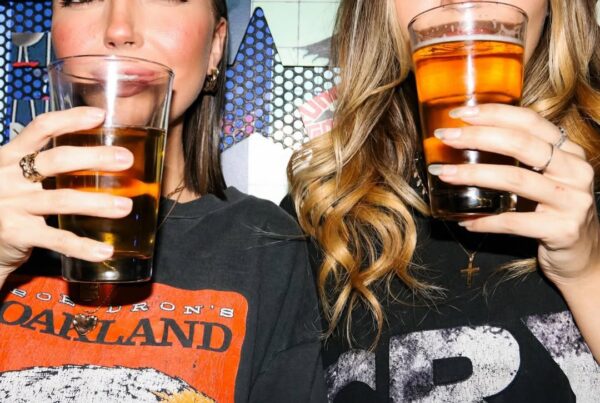 Two women in graphic tees holding up pint glasses of beer with a comic-style mural in the background at Warehouse.
