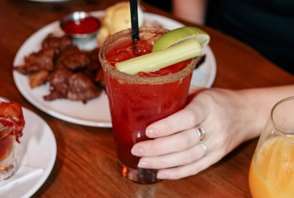 Hand holding a Caesar with celery and lime, surrounded by brunch plates and drinks at Warehouse.