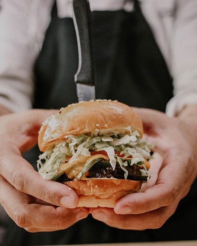 A chef holding a hearty burger layered with a crispy patty, cucumber, pickles, tomatoes, and creamy coleslaw.
