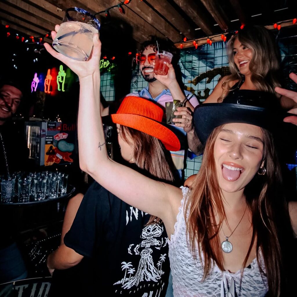 Group of friends partying at Warehouse, raising cocktails with big energy under neon lights and string décor.