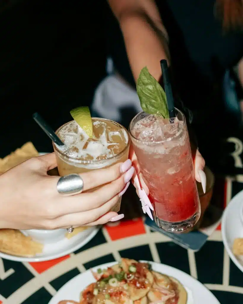 Two colorful cocktails clinking together at Warehouse with plates of food in the background.