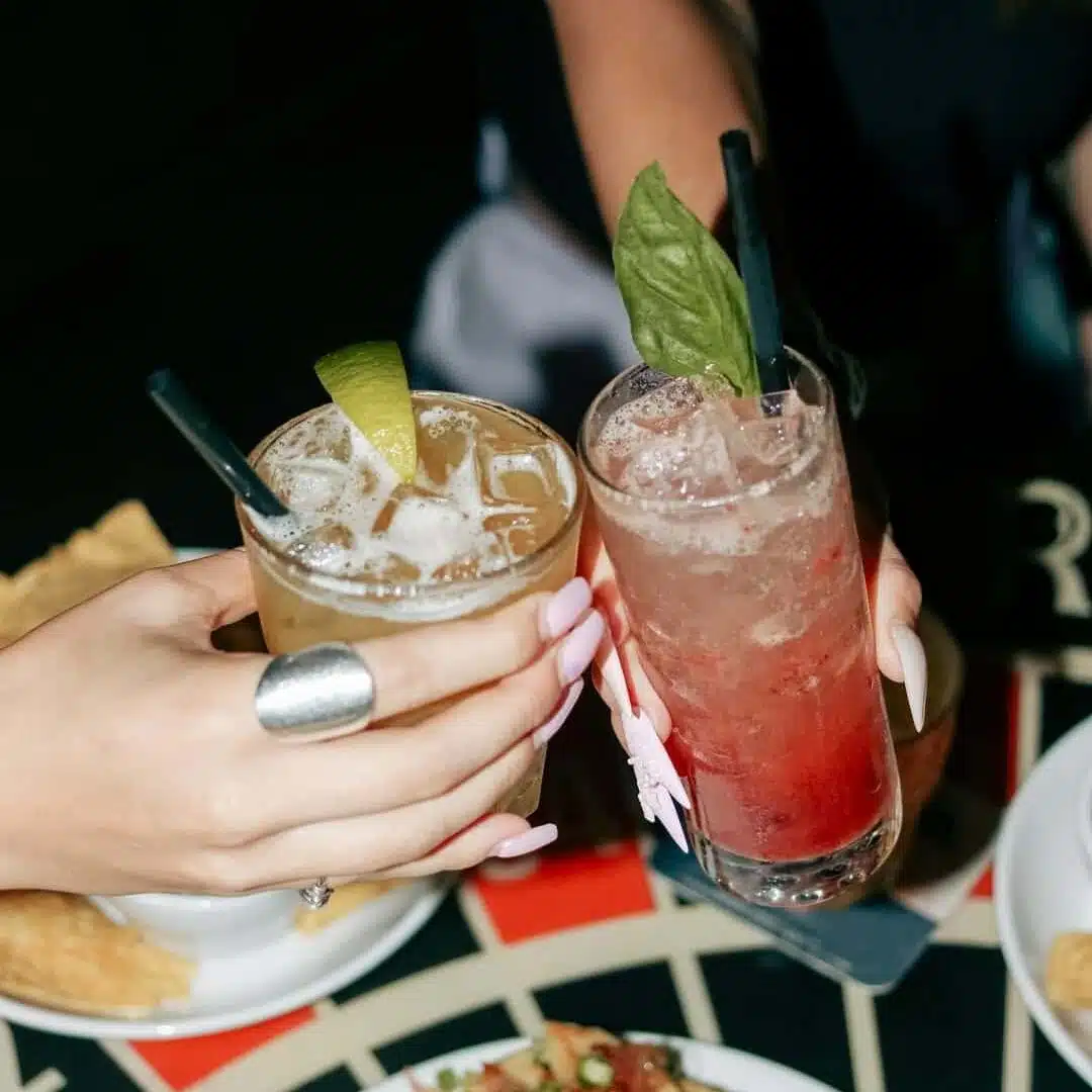 Two people clinking tall glasses of colourful Signature Warehouse cocktails in a lively toast.