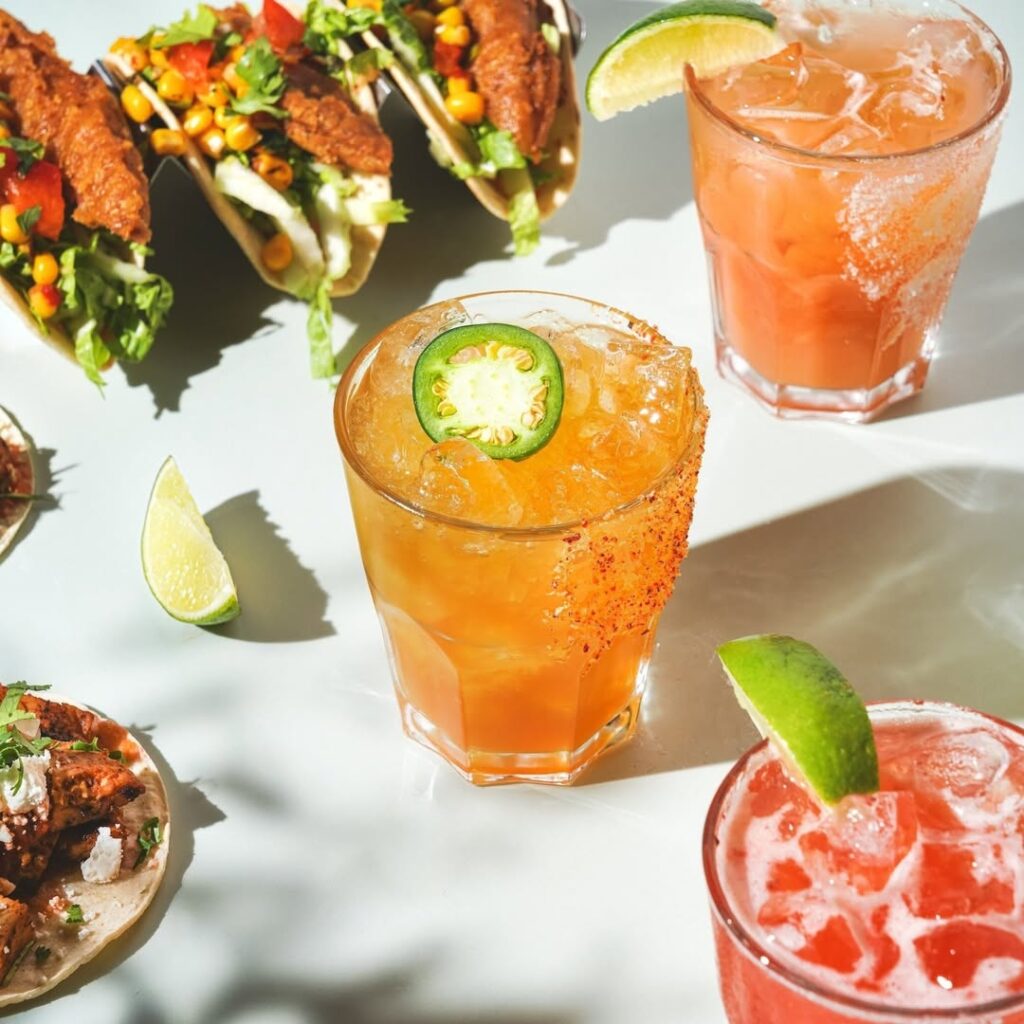 An assortment of Warehouse cocktails, including the Watermelon Jalapeño Mojito, surrounded by street-style soft shell tacos.