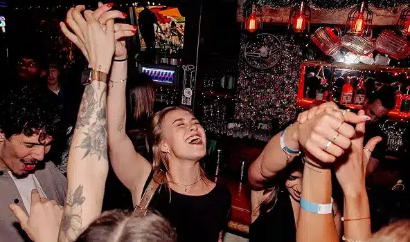 A crowd of people dancing and cheering with their hands in the air at a bar.