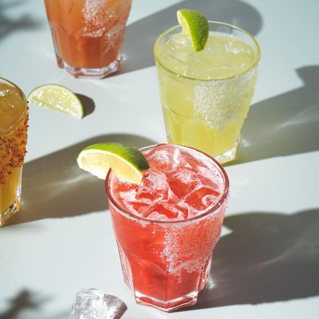 Colourful margaritas and cocktails with salted rims and lime wedges, lit by sunlight on a white tabletop.