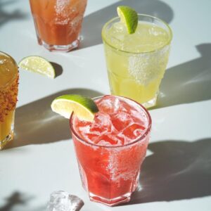 Colourful margaritas and cocktails with salted rims and lime wedges, lit by sunlight on a white tabletop.