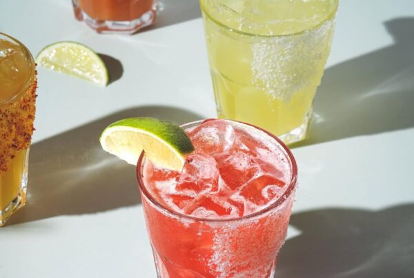 Colourful margaritas and cocktails with salted rims and lime wedges, lit by sunlight on a white tabletop.