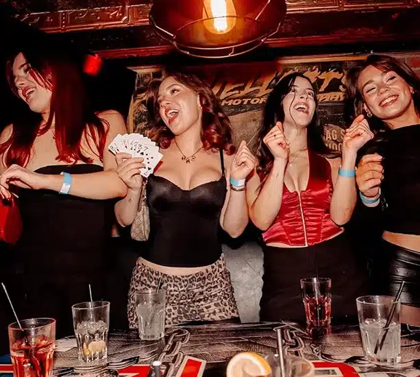 Four women laughing and drinking at a bar, one holding a fan of cards.