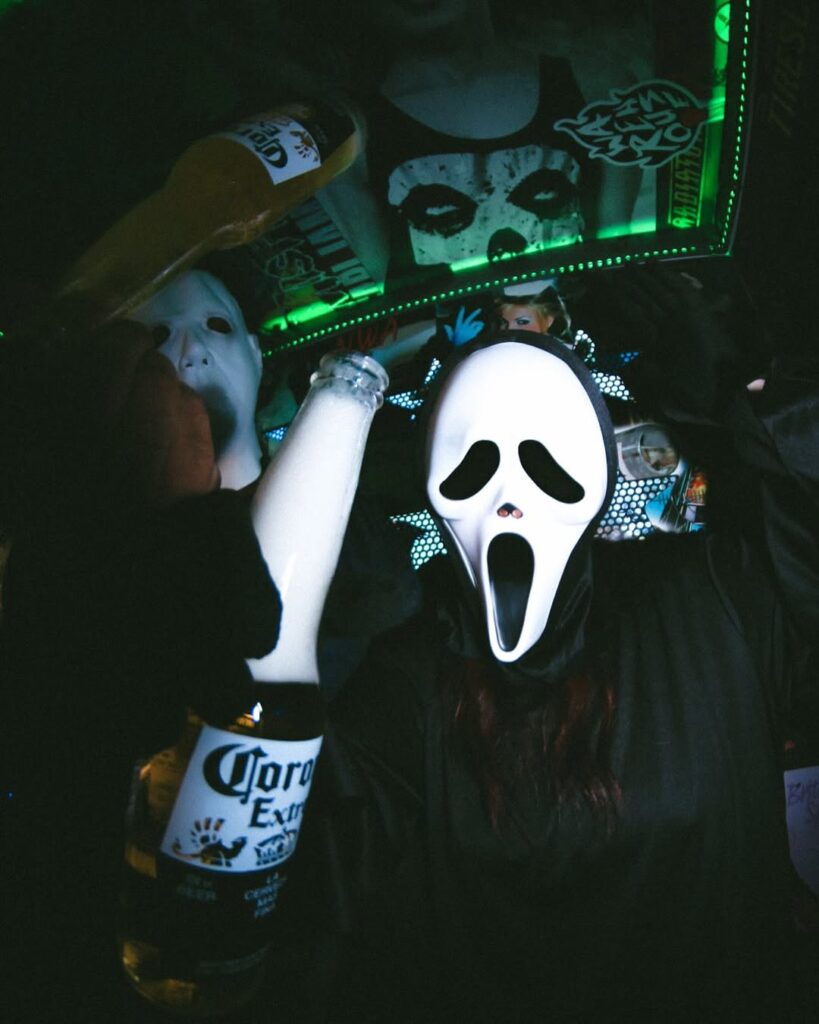 Guests wearing Ghostface masks raising Corona beers during The Warehouse’s Halloween celebration surrounded by party lights.