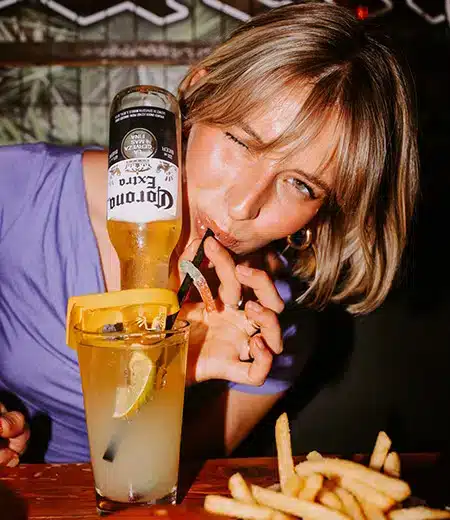 Woman sipping a cocktail topped with a Corona bottle, fries on the table, inside The Warehouse’s lively Halloween setup.
