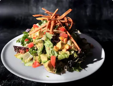 A close-up of a Southwest tortilla salad with crispy tortilla strips, avocado, and corn on a white plate.