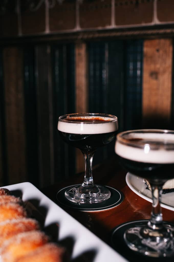 Two espresso martinis served in coupe glasses on branded Warehouse coasters, placed beside a plate of sugar-coated churros.