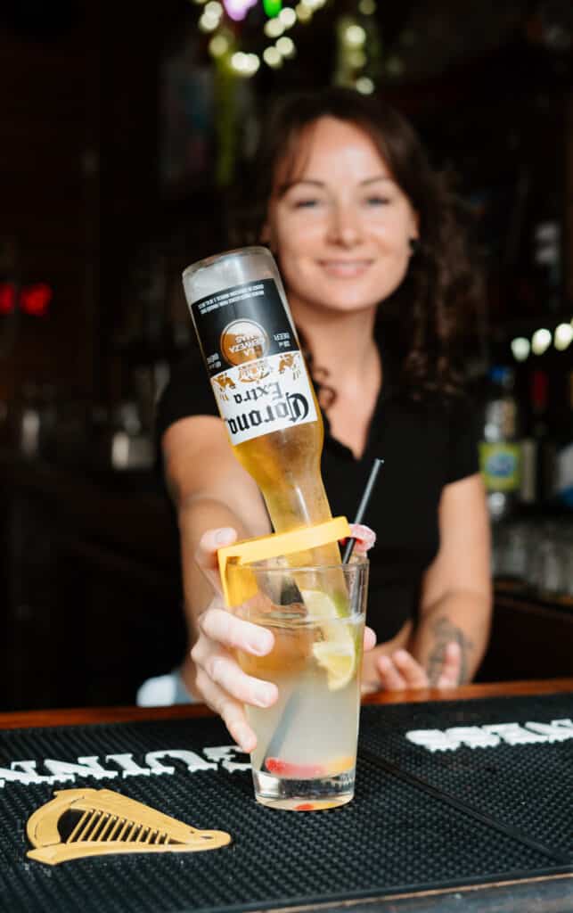 A smiling Warehouse bartender holds a cocktail glass with an upside-down Corona clipped in, ready to serve with flair.
