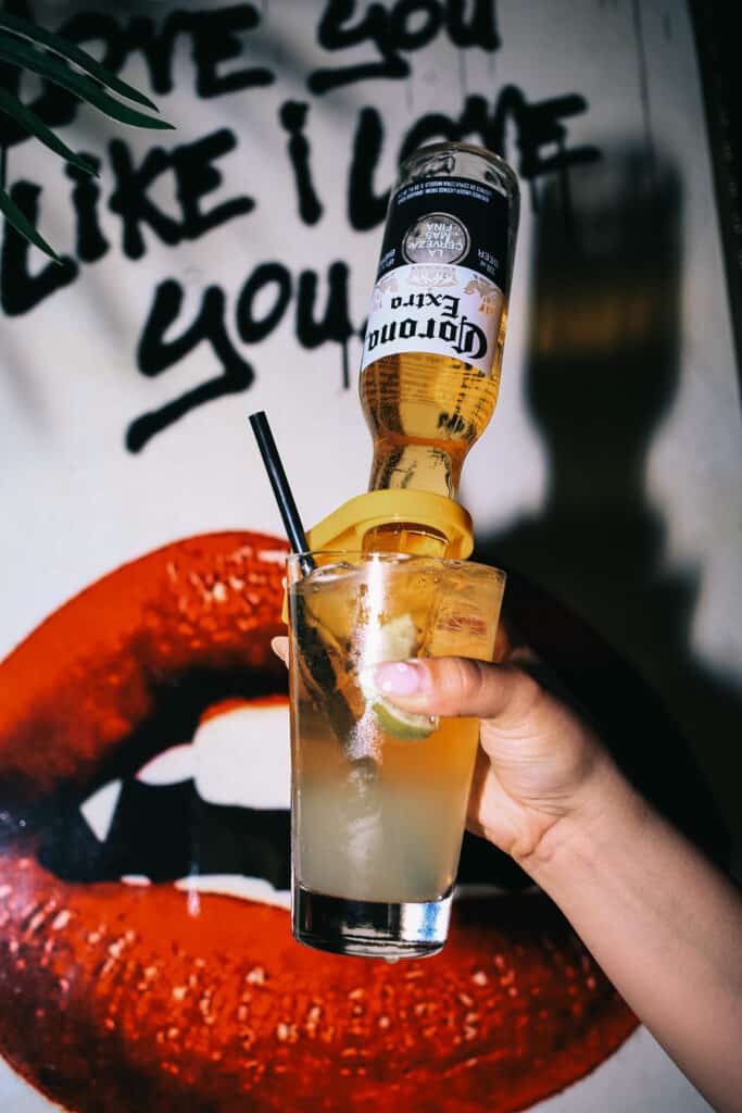A hand holding a Corona beer cocktail in front of a red lips mural and bold script wall at Warehouse.