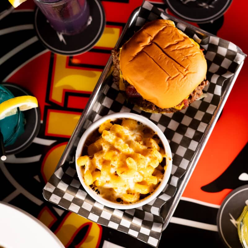 A cheeseburger and a bowl of mac and cheese on a tray, with colorful cocktails in the background.