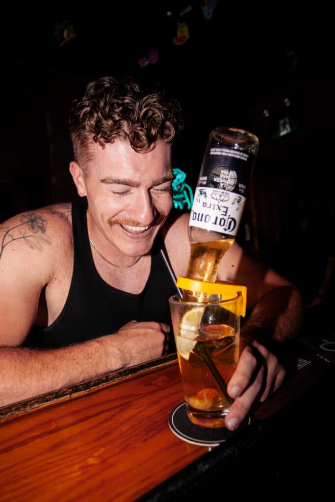 Smiling man at Warehouse bar watching a Corona beer pour into a cocktail glass, capturing a fun, energetic atmosphere.