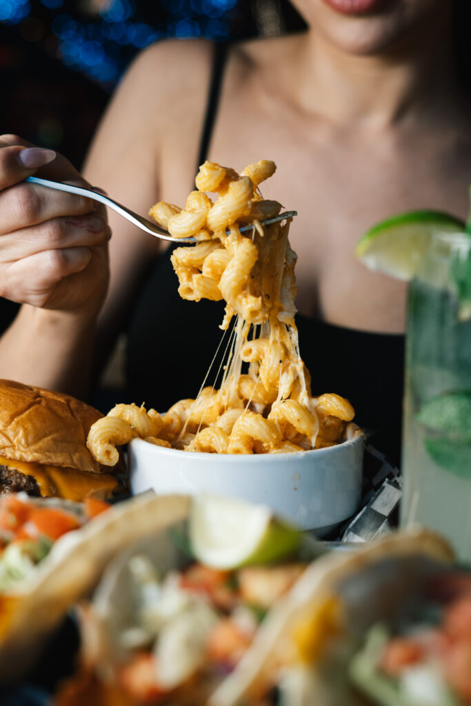 A close-up of someone lifting a forkful of creamy mac and cheese over a bowl, with a burger and tacos in the background.