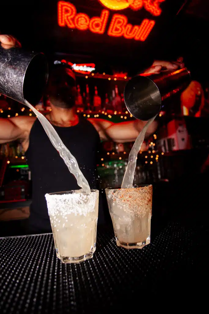 A bartender at Warehouse pours two cocktails simultaneously into salt-rimmed glasses, capturing the vibrant energy of the bar.